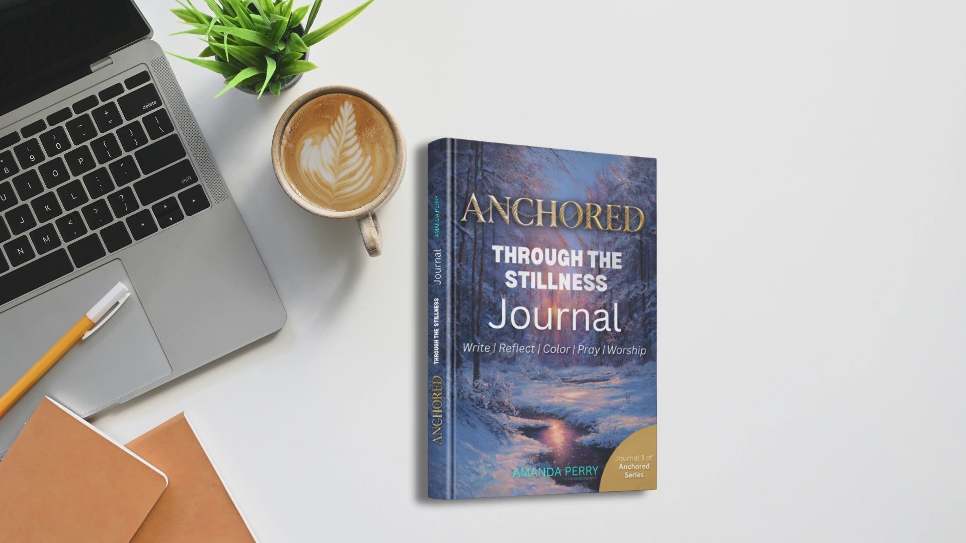 Anchored Through the Stillness book on a desk with laptop. cappucino and pen with desk plant and light background